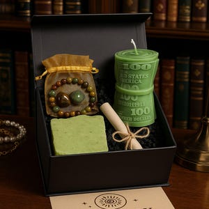 Könnte beinhalten: Eine schwarze Geschenkbox enthält eine grüne Kerze in Form eines Stapels 100-Dollar-Scheine, ein grünes Stück Seife, ein Perlenarmband und eine kleine Schriftrolle, die mit Bindfaden gebunden ist. Eine Karte mit dem Text "AURABOX SHOP" befindet sich im Vordergrund.