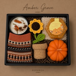 Könnte beinhalten: Schwarze Geschenkbox mit der Aufschrift "Amber Grove", die eine Kerze, Socken, eine Sonnenblume, Seife, einen Kürbis und eine gehäkelte Pflanze enthält. Der Hintergrund ist braun.
