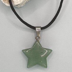 May include: A green star-shaped pendant necklace with a black cord. The star is a light green color and has a silver-colored bail. The necklace is displayed against a light blue background.