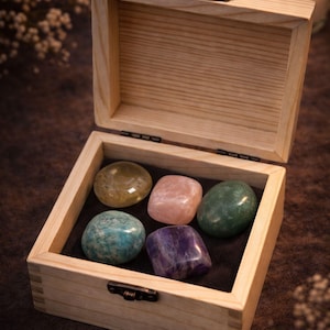 Grief support healing stones set informational cards from AuraBox displayed on a dark warm background with candlelight and dried flowers. The cards explain the meaning of Amethyst, Rose Quartz, Amazonite, Jade and Topaz crystals used for emotional healing, remembrance and comfort during loss. Memorial crystal gift set guide for sympathy, bereavement support and peaceful reflection.