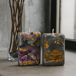 May include: Two rectangular gray candles with gold leaf accents and embedded crystals. One candle features purple and brown crystals, while the other has gold leaf. A clear vase with brown branches is in the background.