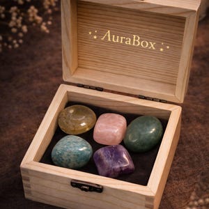 Grief support healing stones set informational cards from AuraBox displayed on a dark warm background with candlelight and dried flowers. The cards explain the meaning of Amethyst, Rose Quartz, Amazonite, Jade and Topaz crystals used for emotional healing, remembrance and comfort during loss. Memorial crystal gift set guide for sympathy, bereavement support and peaceful reflection.