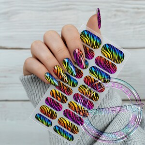 K&ouml;nnte beinhalten: Ein Bogen mit Nagelkunst-Aufklebern mit einem Regenbogen-Zebramuster. Die Aufkleber werden an einer Hand mit passender Nagelkunst gezeigt. Die Nagelkunst-Aufkleber haben ein gl&auml;nzendes Finish und sind in Gelb-, Orange-, Rosa-, Lila- und Blaut&ouml;nen gehalten.