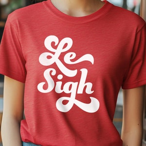 May include: A red t-shirt with the words "Le Sigh" in white, stylized script. The shirt is made of a soft material and has short sleeves. The model is wearing blue jeans.