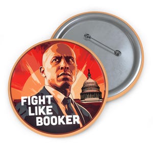 May include: A round button with a red, orange, and yellow background. The image features a man with a shaved head and a serious expression. He is wearing a dark suit and tie. The text "FIGHT LIKE BOOKER" is printed in white on the button. The Capitol Building is visible in the background.