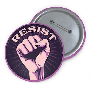 May include: A purple button with a raised fist and the word "RESIST" in white letters. The button has a silver back.