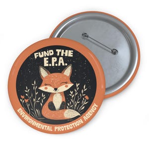 May include: A round button with an orange background and a black circle in the center. The circle features a cartoon fox with a white belly and a black nose, sitting in a field of green grass with red flowers. The text "FUND THE E.P.A. ENVIRONMENTAL PROTECTION AGENCY" is written in a white, semi-circular arc around the fox.