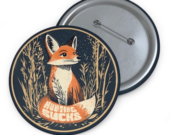May include: A round button with a black background featuring a cartoon fox with an orange and white fur pattern. The fox is sitting in a field of tall grass with the text "Hunting Sucks" printed in orange and white letters on the fox's tail.
