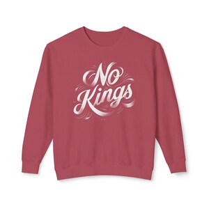 以下が含まれることがあります： マルーン色のスウェットシャツ。白い筆記体で「No Kings」と書かれています。テキストは装飾的な渦巻きと線で装飾されています。スウェットシャツはクルーネックと長袖です。
