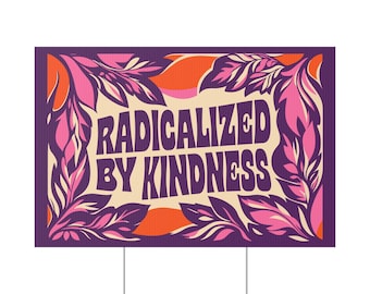 May include: A rectangular yard sign with the text "RADICALIZED BY KINDNESS" in purple and white. The sign is framed by stylized pink and purple leaves and orange accents. The sign is mounted on a metal stake.