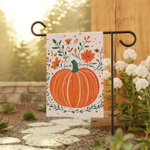 May include: A white garden flag featuring an orange pumpkin illustration with green leaves and flowers. The text "fuck trump" is written above the pumpkin. The flag is displayed on a black metal stand in a garden setting.