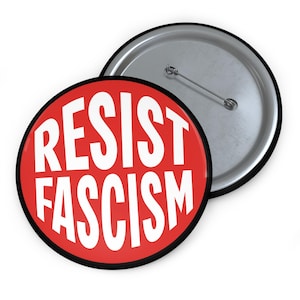 May include: A red button with a black border and white text that reads "RESIST FASCISM". The back of the button is silver.