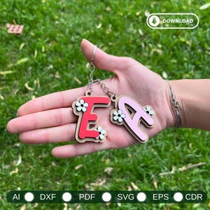 Peut inclure: Deux porte-clés en bois, l'un avec la lettre "E" en rouge et l'autre avec la lettre "A" en rose. Les deux lettres sont décorées d'accents de fleurs blanches et attachées à une chaîne argentée. Les porte-clés sont tenus sur un fond d'herbe verte.