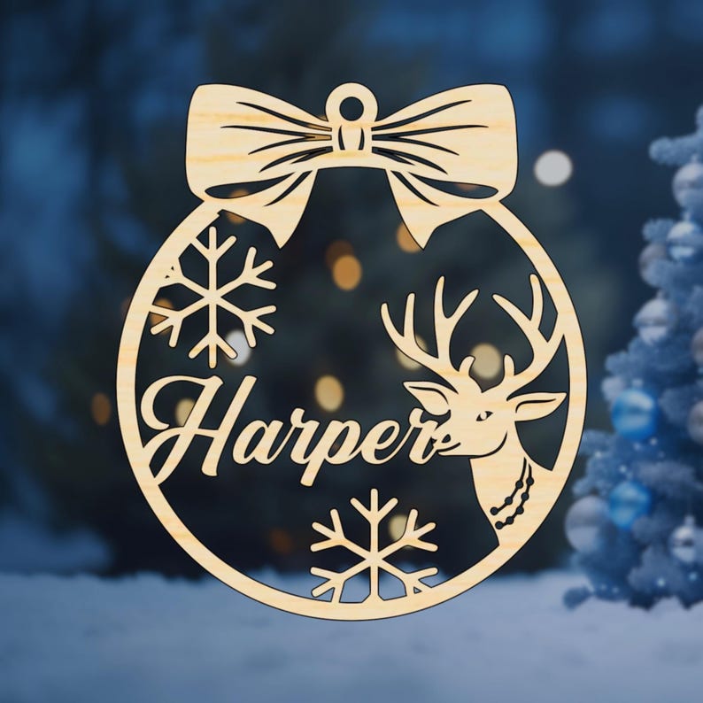 K&ouml;nnte beinhalten: Ein runder Weihnachtsbaumschmuck aus Holz mit einer Schleife oben. Der Schmuck hat einen Hirsch, Schneeflocken und den Namen "Harper", der aus dem Holz geschnitten ist.