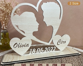 May include: A wooden cutout of two hearts with the names Olivia and Ciro inside. The hearts are joined together and have a date, 18.05.2022, written below them. The cutout is on a wooden base.