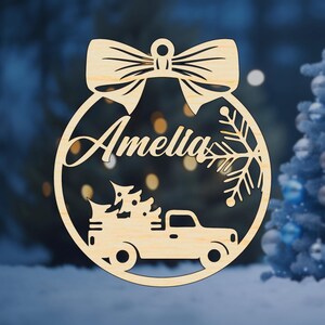K&ouml;nnte beinhalten: Ein runder Weihnachtsbaumschmuck aus Holz mit einer Schleife oben. Der Anh&auml;nger zeigt einen LKW, der einen Weihnachtsbaum transportiert, einen Schneeflock und den Namen "Amelia" darauf.
