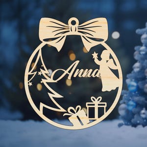 K&ouml;nnte beinhalten: Ein h&ouml;lzerner Weihnachtsbaumschmuck in Form einer Kugel mit einer Schleife oben. Der Schmuck zeigt einen Weihnachtsbaum, einen Engel, zwei Geschenke und den Namen "Anna".
