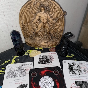 Może przedstawiać: Drewniana rzeźba skrzydlatej postaci z mieczem stoi za kartami tarota. Karty zawierają ilustracje i tekst, w tym "ASMODAY" i "NITH-HAIAH". Ciemny kryształ i inne elementy dekoracyjne są również obecne.