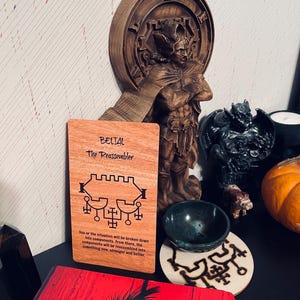 May include: A wooden card with the words "BELTAL The Reassembler" and a symbol printed on it. The card is sitting on a table with a wooden statue and a small bowl.