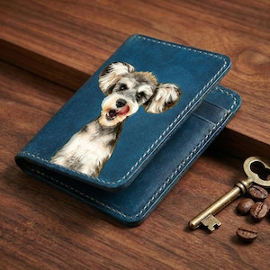 May include: Blue leather wallet featuring a Schnauzer dog portrait. The wallet has white stitching and a bi-fold design. A vintage key and coffee beans are next to the wallet on a wooden surface.
