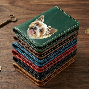 May include: A stack of leather wallets in various colors, including green, brown, blue, and red. The top wallet is green and features a detailed image of a dog's face. The wallets are stacked on a wooden surface.
