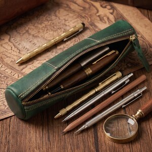 May include: A green leather pen case with a zipper, holding several pens and pencils of various designs and colors. A gold-colored magnifying glass rests on the wooden surface, alongside the writing instruments. The case is approximately 8 inches long.