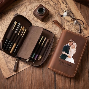 Op de afbeelding: Een bruine leren etui met meerdere pennen en een bijpassende portemonnee met ritssluiting. De portemonnee toont een foto van een stel met de namen "Tyler & Sofia" en de datum "02/08/2026". Een vintage kaart en een inktpot zijn ook zichtbaar.