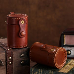 May include: Two brown leather cylindrical cases with brass hardware. One case stands upright, while the other lies on its side. Both have a stitched design and a flap closure. The cases are on a wooden surface.