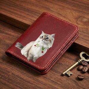 May include: A burgundy leather wallet featuring a detailed illustration of a fluffy cat wearing a festive collar. The wallet is rectangular with stitched edges. A vintage key and coffee beans are next to the wallet on a wooden surface.
