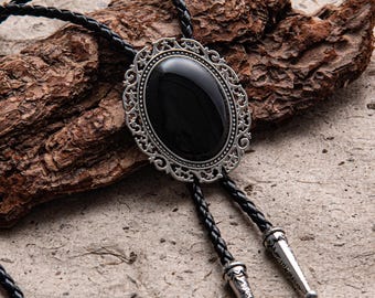 May include: A black bolo tie with a silver-colored filigree oval pendant and a black braided leather cord. The pendant features a large, polished black stone. The cord ends are silver-toned with decorative detailing.
