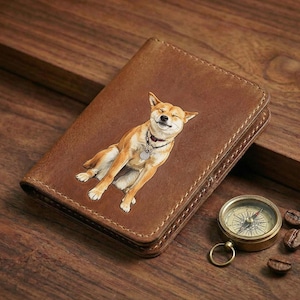 May include: A brown leather wallet with a smiling Shiba Inu dog illustration. The wallet is rectangular and has a stitched border. A small brass compass and coffee beans are next to the wallet on a wooden surface.