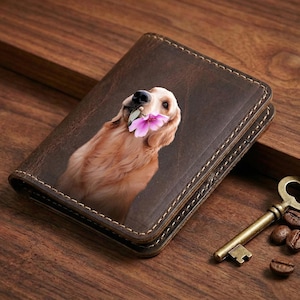 May include: A brown leather wallet featuring a golden retriever holding a pink flower in its mouth. The wallet has a stitched border and a vintage aesthetic. A bronze key and coffee beans are next to the wallet.