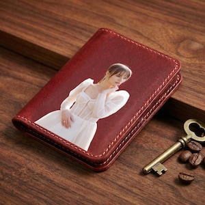 May include: A burgundy leather wallet with a photograph of a woman in a white dress. The wallet is rectangular with stitched edges. A vintage key and coffee beans are next to the wallet on a wooden surface.