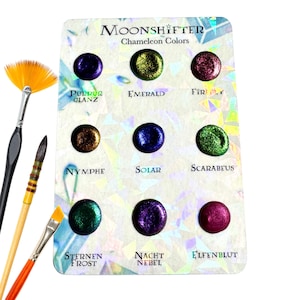 May include: A Moonshifter Chameleon Colors paint palette featuring nine circular swatches of iridescent pigment in shades of purple, green, gold, and red, with labels including Purpur Glanz, Emerald, Nymphe, Solar, Scarabeus, Sternen Frost, Nacht Nebel, and Elfenblut.