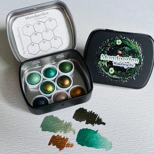 Könnte beinhalten: Eine schwarze Metalldose mit sieben kleinen runden Aquarellnäpfchen in Grün-, Bronze- und Dunkelblautönen. Der Deckel zeigt ein Waldmotiv und den Text Mondperlen Waldmystic. Im Deckel befindet sich eine Farbkarte mit nummerierten Sechsecken.