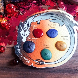 May include: A watercolor palette with a dragon design. The palette has six paint wells labeled "Thorn Red", "Shruikian Black", "Firnen Green", "Glaedr Gold", "Sapphire Blue", and "Dragons Nest".