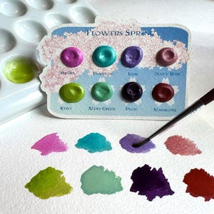 May include: A watercolor paint set with eight metallic colors, including Sakura, Phantom, and Lilac, displayed on a white palette. The card reads "FLOWERS SPRING". A paintbrush is positioned near the paint samples.