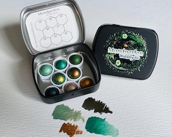 May include: A black metal tin containing eight round watercolor pans in shades of green, gold, and brown. The lid features the text Mondperlen Waldmystic by Moonshineflowers. A color chart is inside the lid, and four paint swatches are below.