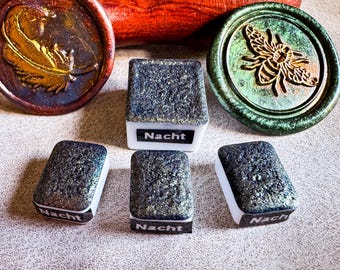 May include: An assortment of decorative wax seals and rectangular blocks. The seals feature embossed designs of a feather and a bee. The blocks have a textured, glittery surface and the word "Nacht" printed on them.