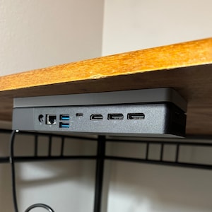 May include: A black computer device with multiple ports is mounted under a wooden desk.