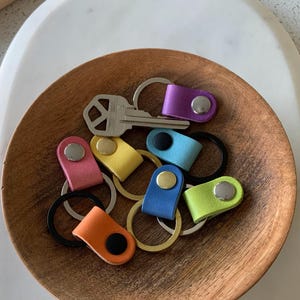 May include: A collection of colorful keychains in a wooden bowl. The keychains feature leather straps in various colors, including purple, pink, yellow, blue, orange, and green, each with a metal rivet. A silver key is also present.