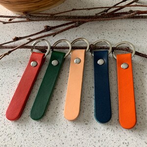 May include: Five leather keychains in a row, each with a silver metal ring. The keychains are in various colors: red, green, peach, navy blue, and orange. Each keychain has a silver metal rivet.