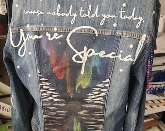 Könnte beinhalten: Eine blaue Jeansjacke mit dem weißen Schriftzug "In case nobody told you today, You're Special". Auf der Rückseite befindet sich ein Gemälde der bunten Aurora Borealis über Wasser, verziert mit Perlen.