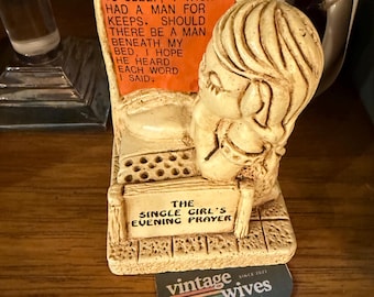 May include: A beige resin figurine of a person kneeling in prayer beside a bed. An orange plaque reads: NOW I LAY ME DOWN TO SLEEP, I WISH I HAD A MAN FOR KEEPS. SHOULD THERE BE A MAN BENEATH MY BED, I HOPE HE HEARD EACH WORD I SAID. A sign reads: THE SINGLE GIRL'S EVENING PRAYER.