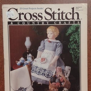 Può includere: La copertina della rivista "Cross Stitch & Country Crafts" presenta una bambola con un vestito a quadri blu e bianco e un grembiule. Sono visibili anche un cesto di palline colorate, un cagnolino e oggetti a punto croce.