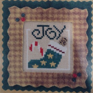 May include: A cross-stitch design featuring a green stocking with white stars and the word "Joy" in a cursive font. The stocking is surrounded by a brown frame with a green border.