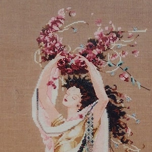 Könnte beinhalten: Eine Kreuzstich-Stickerei einer Frau mit langen braunen Haaren, die ein weißes und goldenes Kleid trägt und einen Strauß rosa Blumen über ihrem Kopf hält.