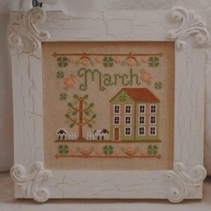Op de afbeelding: Een ingelijst kruissteekkunstwerk met een crèmekleurige achtergrond. Het kunstwerk toont een huis, een boom, schapen en het woord "March". De witte lijst heeft sierlijke hoekdetails. Het huis heeft een groene gevel en een rood dak.