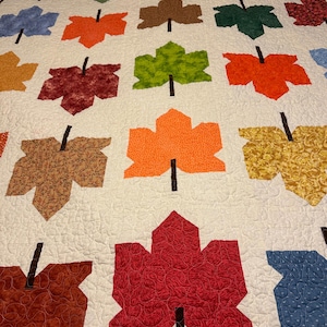 May include: A quilted blanket featuring a repeating pattern of stylized maple leaves in various fall colors, including red, orange, yellow, green, and brown. The leaves are set against a cream-colored background with quilted stitching.
