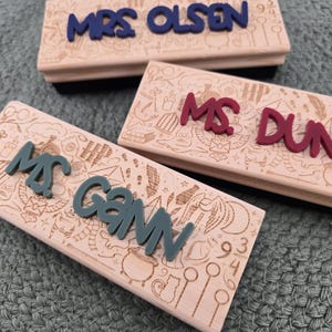 May include: Three wooden stamps with a Harry Potter theme. The stamps are engraved with the names "Mrs. Olsen", "Ms. Dunn", and "Mrs. Gann". The names are in different colors, including red, blue, and green.